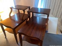 Image showing two dark brown wooden end tables with rectangular tabletops, curved supports between two levels, and straight legs on carpeted floor near window.