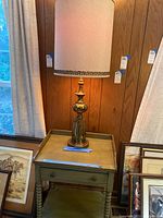 Full view of brass base lamp with large fabric shade sitting on wooden side table with spindle legs and drawer.