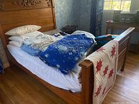 Wooden bed with pillows, blue floral comforter, baby quilt, sheets and blankets on top; blue-patterned curtains in window behind bed.