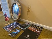 Front view showing marine mirror leaning against wall with eagle ornament atop, small American flag, binoculars, Casio watch and travel clock in packages, and marine books arranged on floor.