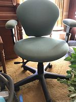 Front view of office chair showing green padded seat, backrest, and armrests, black base with wheels, and adjustment lever under seat.