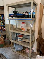 Full view of plastic shelving unit fully assembled and standing against a white brick wall in a garage, showing five shelves with various items placed on them (contents not included in sale).