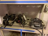 Three vintage power tools including a corded Black & Decker drill (left), a Craftsman jigsaw (middle), and a Craftsman large belt sander (right) on a metal shelf.