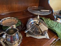Lot contents showing glass cake stand with metal base, candle snuffer, cake slicer with textured handle, metal divided serving dish with serving utensils, and additional metal serving items