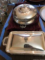 Photo showing oval silverplate casserole dish with lid featuring floral engravings and decorative feet and handles, along with rectangular silverplate covered dish with polished surface and ornate handles.