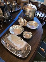 Photo showing various silverplate serving pieces on wooden table including lidded ice bucket, coffee pots, gravy boat, butter dish, bowls, and tray.