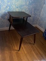 Two-tier wooden end table positioned against a blue textured wall on a hardwood floor, showing the overall shape and structure.