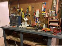 Wide angle photo showing gray workbench with mounted power stapler, red vice, miter saw, various small tools and hardware on and under bench, with pegboard on wall holding hand tools and accessories.