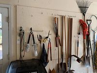 Full view of pegboard wall with various hanging garden and hand tools along with an electric weed trimmer and rakes.