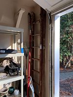 Wooden extension ladder standing vertically against the wall, showing wooden rails and rungs with signs of use and age. Next to shelving and garden tools.