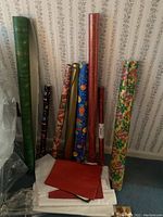 Eight assorted rolls of wrapping paper leaning against a wall with some tissue paper sheets on the floor