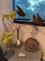 Photo of faux floral plant with large green leaves in clear glass vase, metal-covered decorative storage box, and decorative bird nest in acrylic holder.