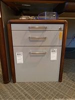 Metal file drawer unit with three drawers and lock, wood trim sides, located under a desk on carpet flooring.