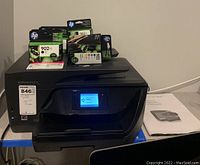 Front view showing printer with touchscreen display, ink cartridge boxes on top, manual at side