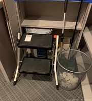 Wide view of shelving with paint bucket on bottom shelf, Polder two-step stool positioned in front, Hoky Oreck sweeper leaning in corner, metal wire trash bin with paper waste on floor next to stool.