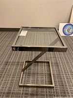 Side angled view of square metal framed side table with glass top on a carpeted floor.