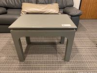 Front view of the painted side table showing gray top and wood legs, set against a carpeted floor and dark leather couch.