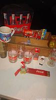 Wooden Pepsi crate with visible wear, Coca Cola metal cooler box, assorted Coca Cola branded glassware, mug, toy truck, tin can, pencils, and ornaments