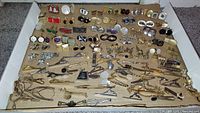 Photo showing several rows of cufflinks and tie clips arranged in a box on a beige base, with a variety of metal finishes and designs.