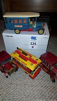 Photo showing four tin toy vehicles: large blue Autobus on top of box, yellow and red Broadway Trolley, and two maroon jalopies on carpet surface.