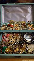 Top view inside vintage blue jewelry box showing assorted pins, earrings, brooches, and colorful costume jewelry pieces in two compartments.