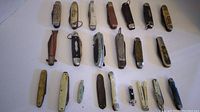Full spread of 23 assorted pen and pocket cutters with various handle materials and sizes, some with keyrings, laid on white background.