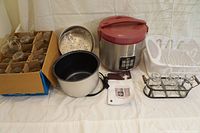 Wolfgang Puck slow cooker with accessories including inner pot, metal steamer basket, instruction manual, small white dish rack and a tray with glass small jars in metal frame.