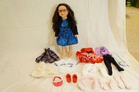 Full view of doll standing with spread clothing and accessories laid out in front on white background.