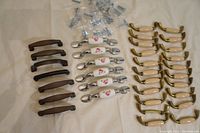 Overview of all cabinet door and drawer knobs and handles arranged in groups on a table, including bronze handles, white rose-patterned ceramic handles, gold curved handles, and silver oval knobs, screws included.