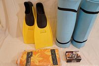 Photo showing yellow fins, two rolled blue sleeping mats, packaged orange floral tablecloth, and boxed Tasco binoculars.