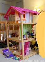Front view of wooden dollhouse with pink roof, showing open sides, furniture pieces including bed, tables, chairs, and accessories on floor.