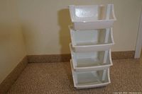 Four white plastic stackable bins stacked vertically on carpeted floor near wall.