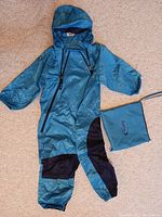 Blue one-piece child rain suit with hood and front zipper, laid flat on carpet with matching pouch beside it