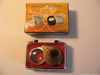 Photo of jeweler's loupe in clear plastic case with red interior and original packaging box shown above it. The packaging is orange with images of different loupe models and labeled 'JEWELER’S LOUPE'.