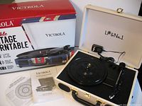 Turntable set up in open white suitcase with black platter and tonearm. Comes with power adapter and instruction manual. Original box in background.