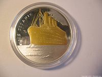 Obverse side of silver and gold plated coin showing RMS Titanic ship in gold plating floating on water with detailed silver background and dotted border, in original plastic capsule.