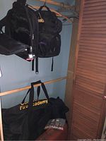 Photo showing two black backpacks hanging side by side on hangers, with a black duffel bag on floor below.