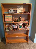 Front view of wooden bookcase with five shelves, containing various items including books, plush toys, model, and small decorative items. Shelf surfaces show wear and use.