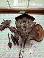 Overall view of cuckoo clock and detached parts on patterned cloth, showing carved wood design, chains, weights, pendulum, and clock face
