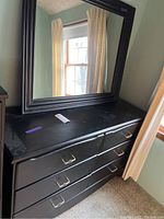 Full view of the black wooden dresser with six drawers and silver handles, showing scratches on the top surface and part of the matching mirror frame above it.