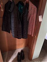 Three jackets and boots hanging inside closet. Visible are two black puffer vests and one red-white-black checked jacket with boots below them.