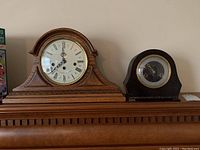 Two mantle clocks side by side: brown Howard Miller 60th Anniversary wooden clock and black Smiths Enfield clock.