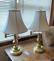 Pair of gold-colored metal base table lamps with cream fabric shades on wooden table, with window blinds in background.