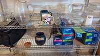 Various glass pitchers, mugs, vases, glass storage containers with lids, and boxes of Ziploc bags on metal shelving.