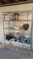 Full view of metal wire storage shelf unit containing various household items such as a Cuisinart food processor box, kitchen bowls, mugs, plastic storage bags, and other kitchen containers.