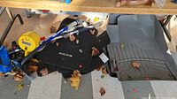 View of two black KICKS carpet mats, WeatherTech winter rubber mats, yellow measuring tape, car washing scrub brushes, and cleaning cloth placed on garage floor under a table, with some dry leaves on them.