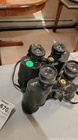 Photo of two black Bushnell binoculars with brass knobs and neck straps shown on a grey surface.