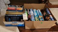 Two cardboard boxes filled with Clive Cussler books, some hardcover with dust jackets and some softcover, arranged standing upright and stacked.