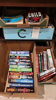 Four boxes of softcover books stacked on shelves and floor showing Clive Cussler novels prominently