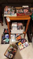Photo showing multiple DVDs, CDs, and books arranged on and around a small table. DVDs are in various packaging formats including boxes and jewel cases. CDs are in standard and paper sleeves, some loose. Books partially visible.